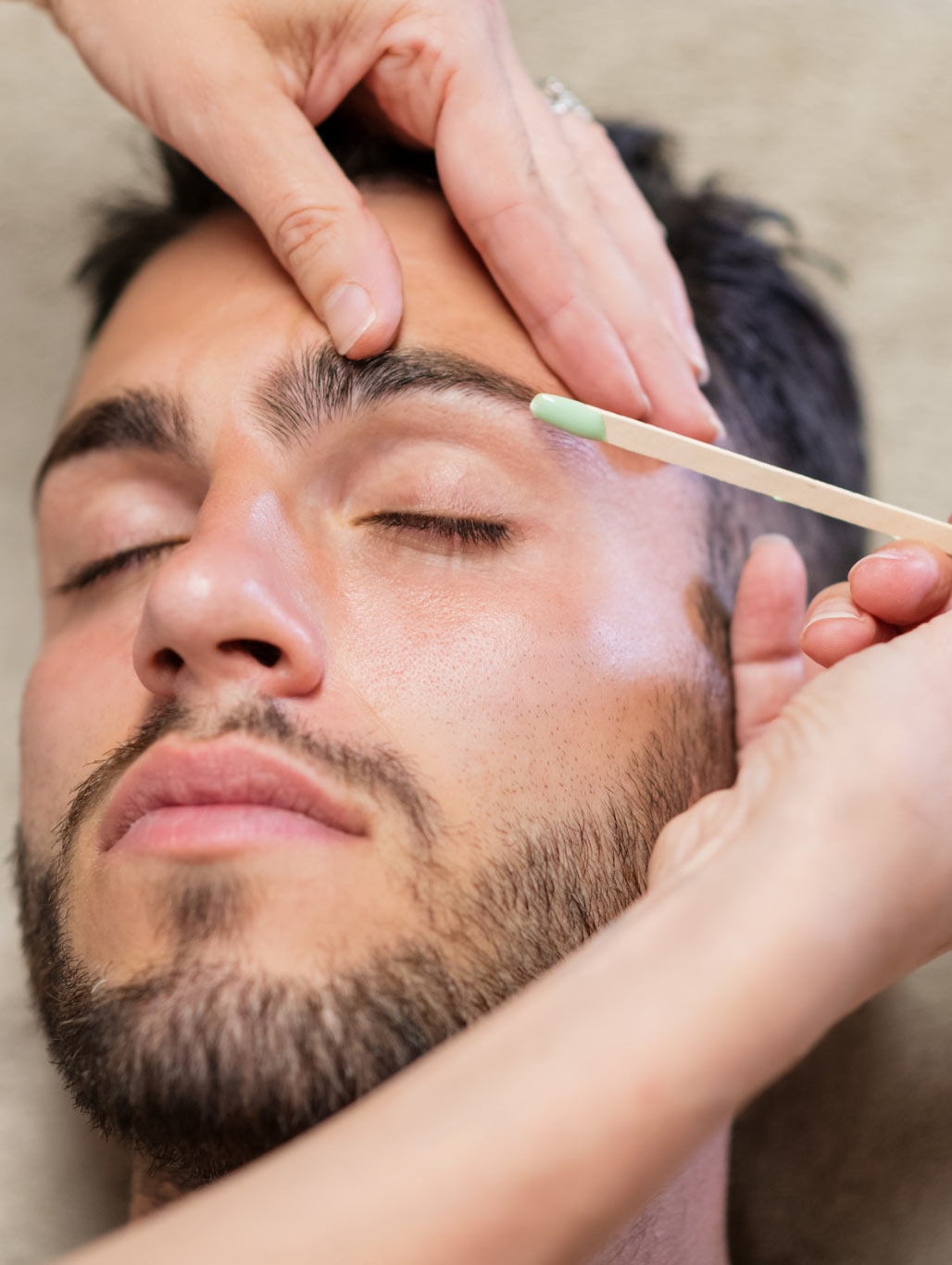 Close-up of a person with closed eyes getting eyebrows waxed by another person's hands using a wooden stick.