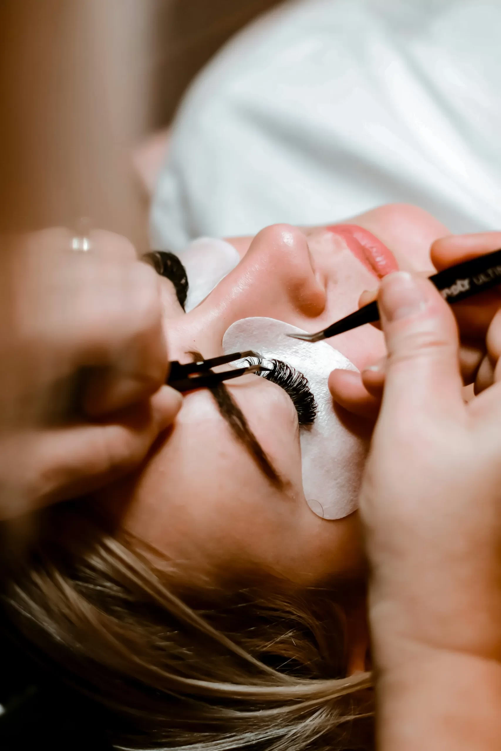Close-up of a person receiving eyelash extensions, with hands applying lashes and eye pads under the eyes.